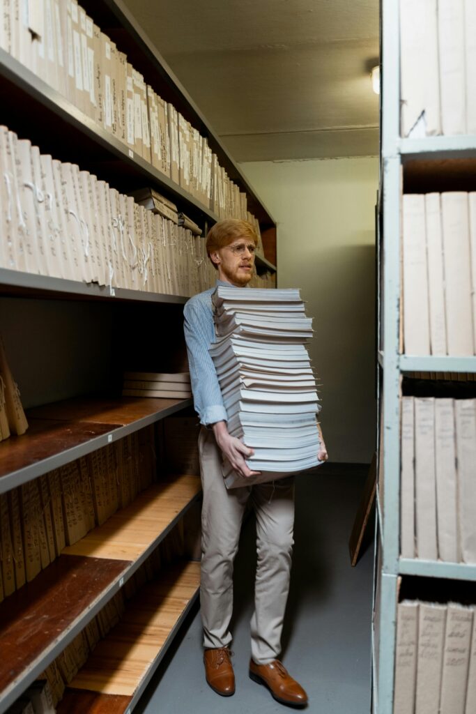 pexels photo 8872373 8872373 Adult man in archive room carrying a large stack of documents, surrounded by shelves.